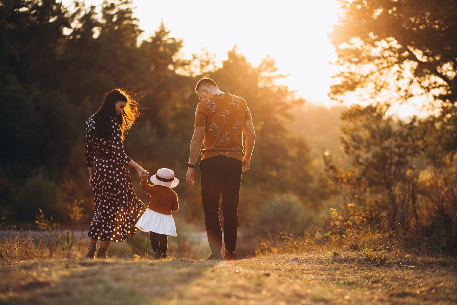 Parents walking a path at sunset holding the hands of their small daughter in a sun hat