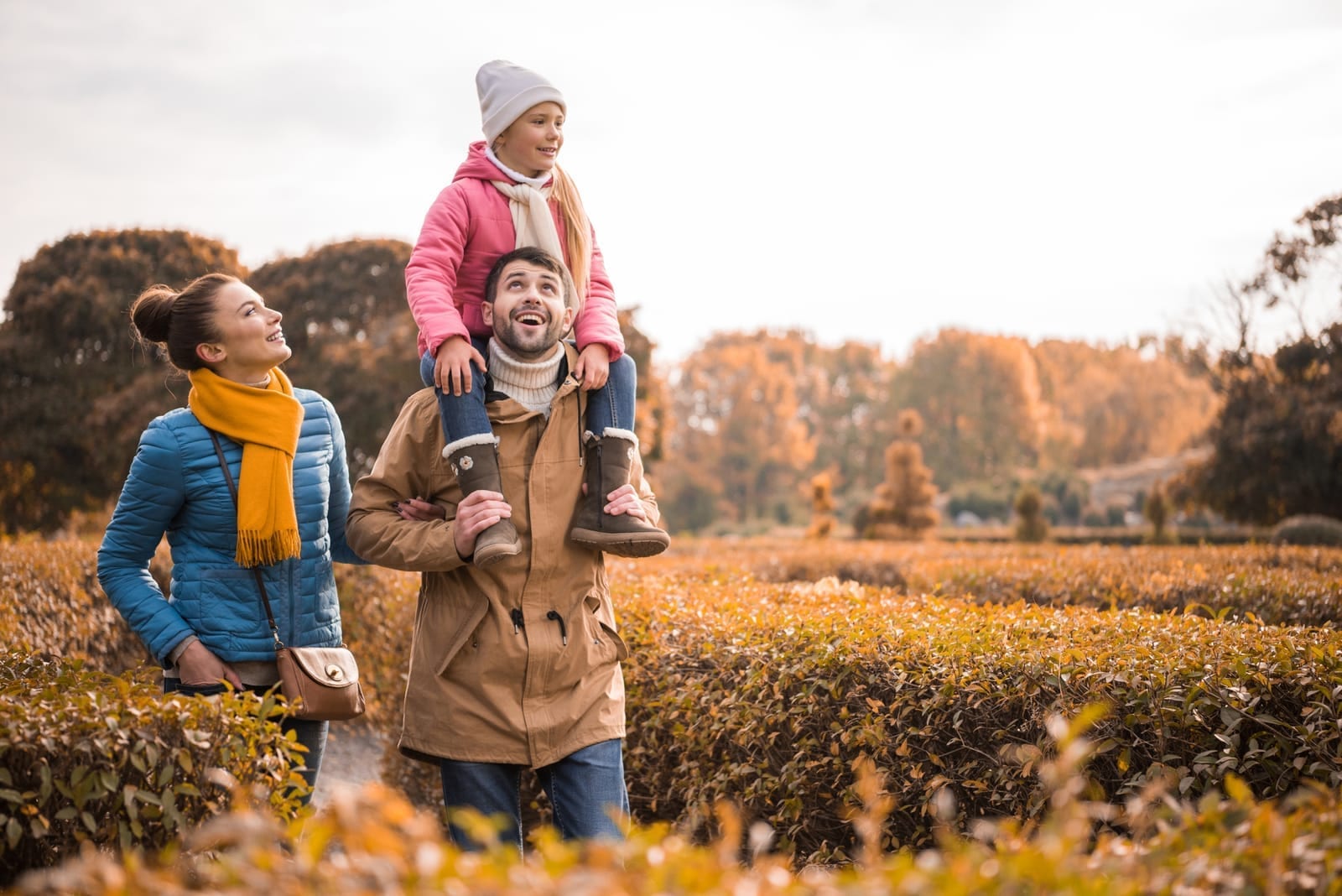 A mother, father and young daughter walking through an autumn park, the girl riding on her father's shoulders