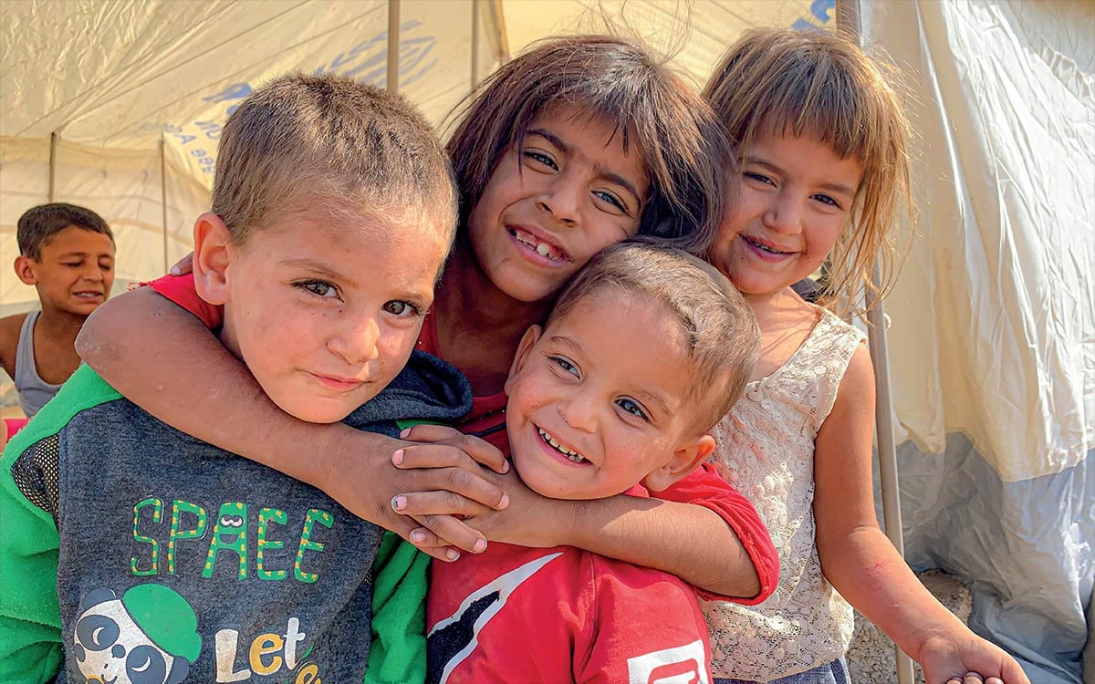 Four Syrian refugee children stand with their arms round each other outside a canvas tent, smiling at the camera, another boy behind them.