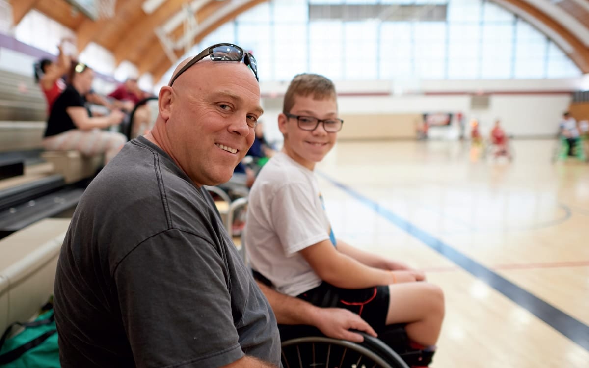 A man sitting courtside in a school gym turns to smile at the camera beside a boy in a wheelchair, a wheelchair basketball game going on behind them.