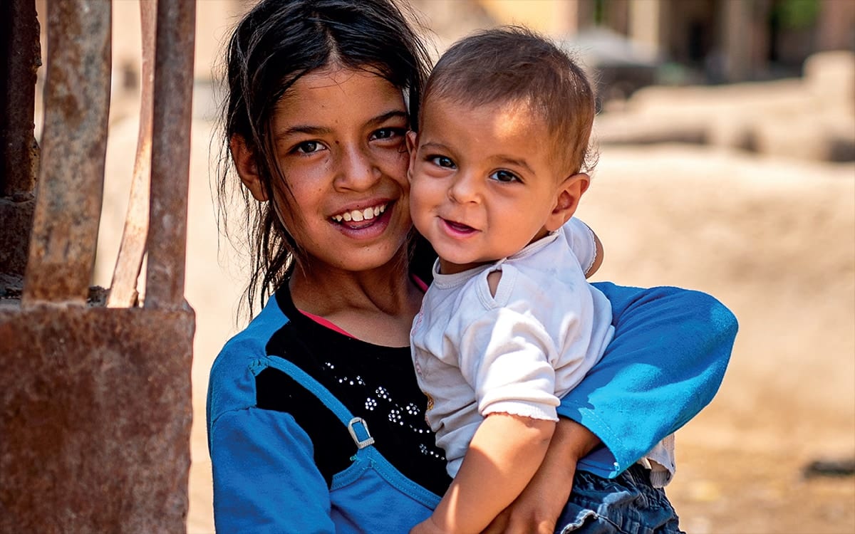 A girl carries a baby on her hip in a dusty street, both of them smiling at the camera.