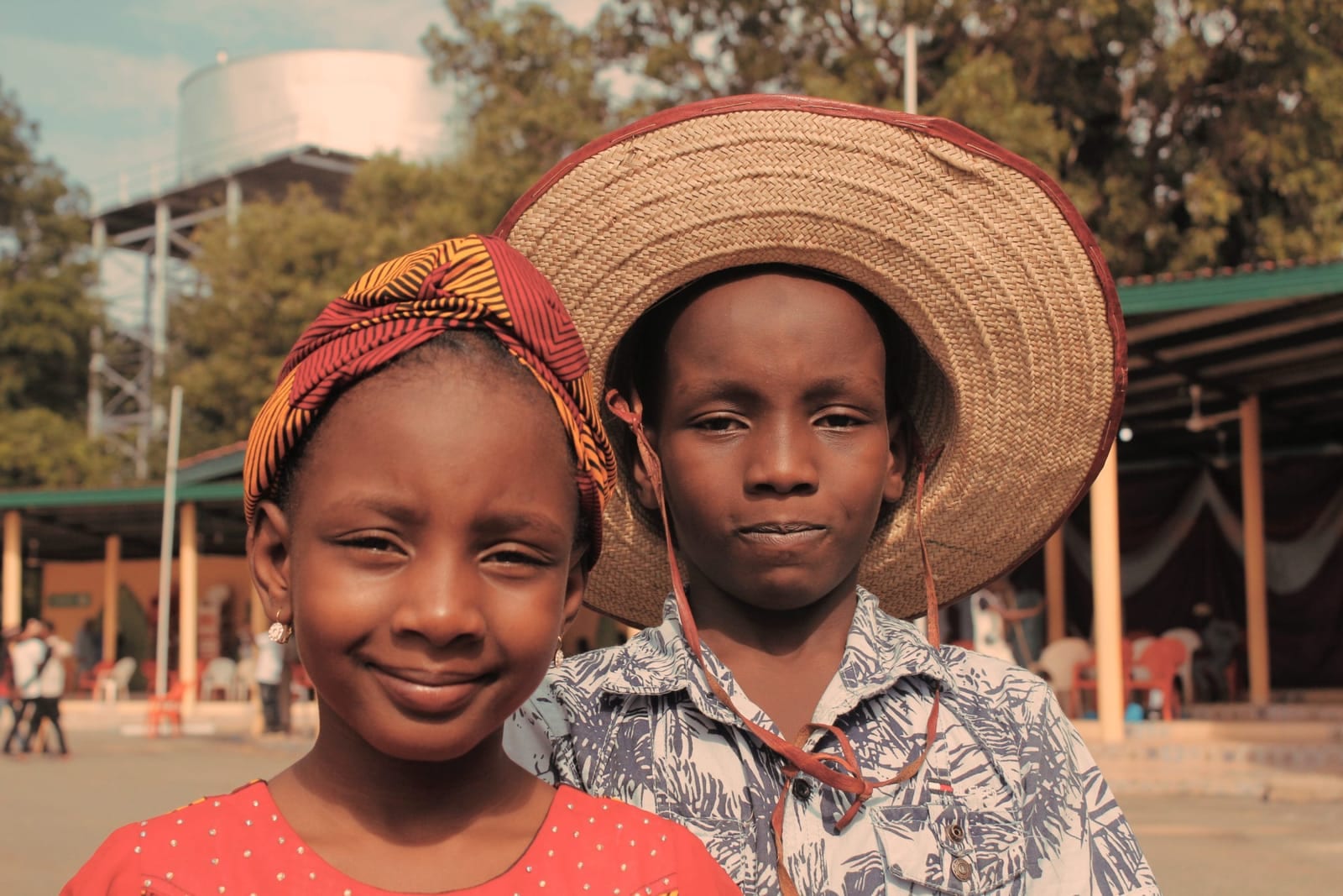 A girl in a patterned headwrap and a boy in a wide straw hat standing side by side outdoors, looking at the camera