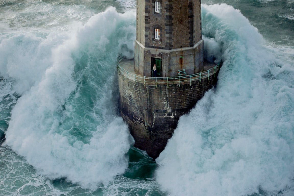 A lighthouse in a heavy sea, a huge wave breaking around its base, a person standing in the open doorway