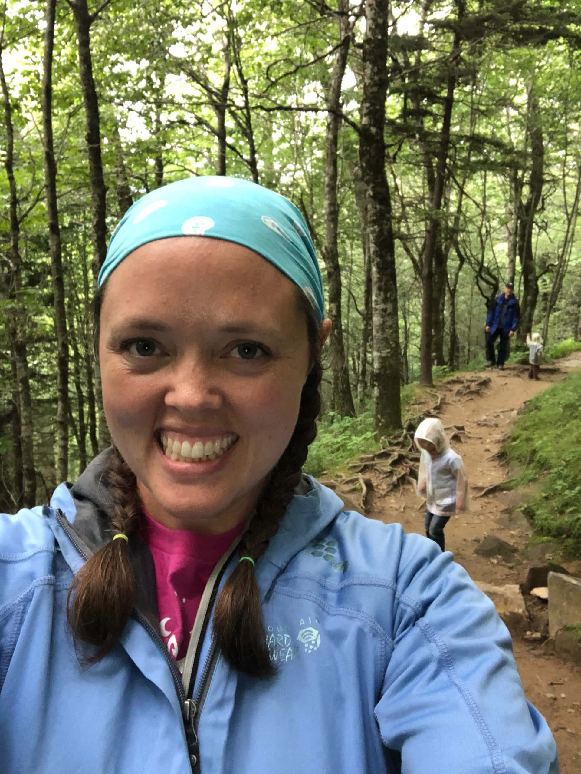 A woman smiling at the camera on a wooded mountain trail, in a bright headband and rain jacket, with a small child in a white rain poncho and two more of her family climbing the path behind her.