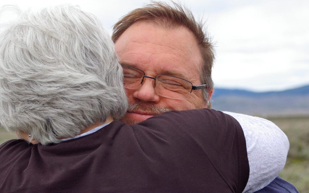 An older woman with white hair holds a middle-aged man in a long embrace, his eyes closed and his face resting against her shoulder, on open ground with hills behind them.