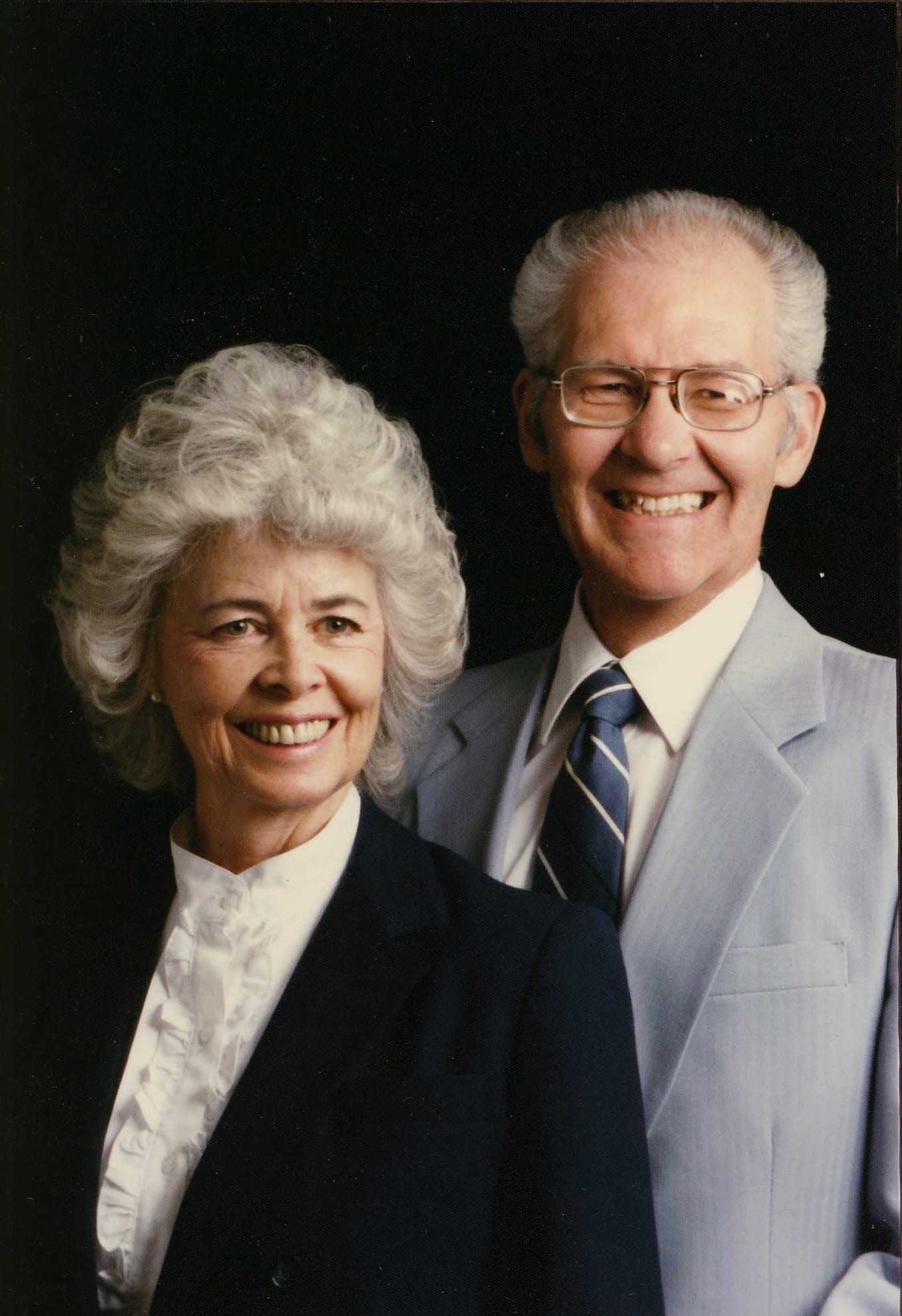 A studio portrait of an older couple against a black background, both smiling, she in a dark jacket over a white blouse and he in a light grey suit and striped tie.